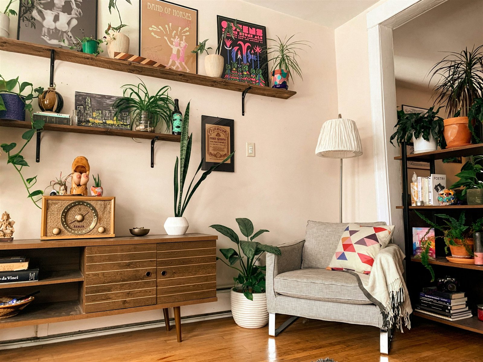 Esquina de salón con estantes de madera, pósters enmarcados, plantas y un sillón gris con cojín geométrico